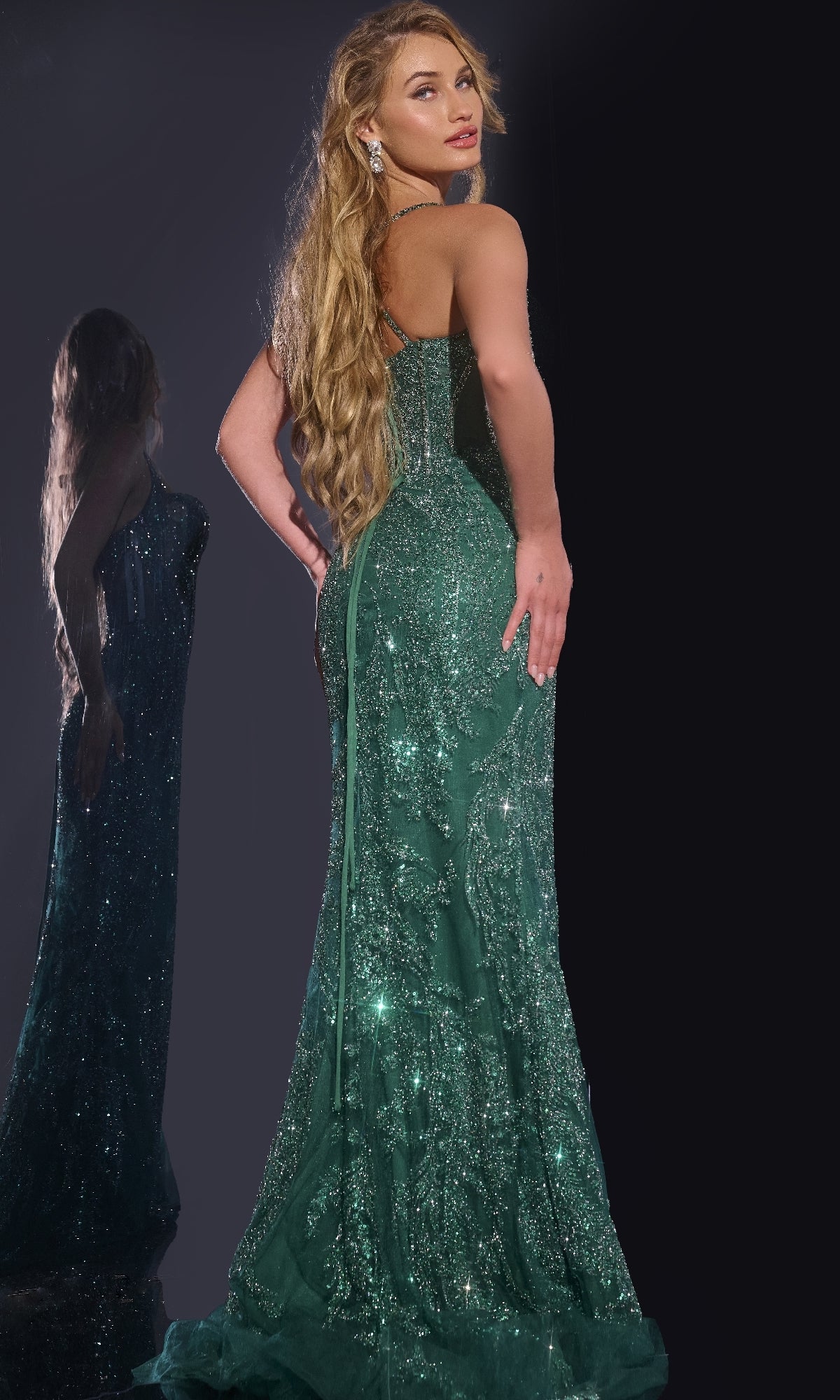 Woman with long blonde hair wears the Jovani 40470 Lace-Up Long Beaded Prom Dress in sparkling green, posing with her back turned and looking over her shoulder; her reflection appears on the left.